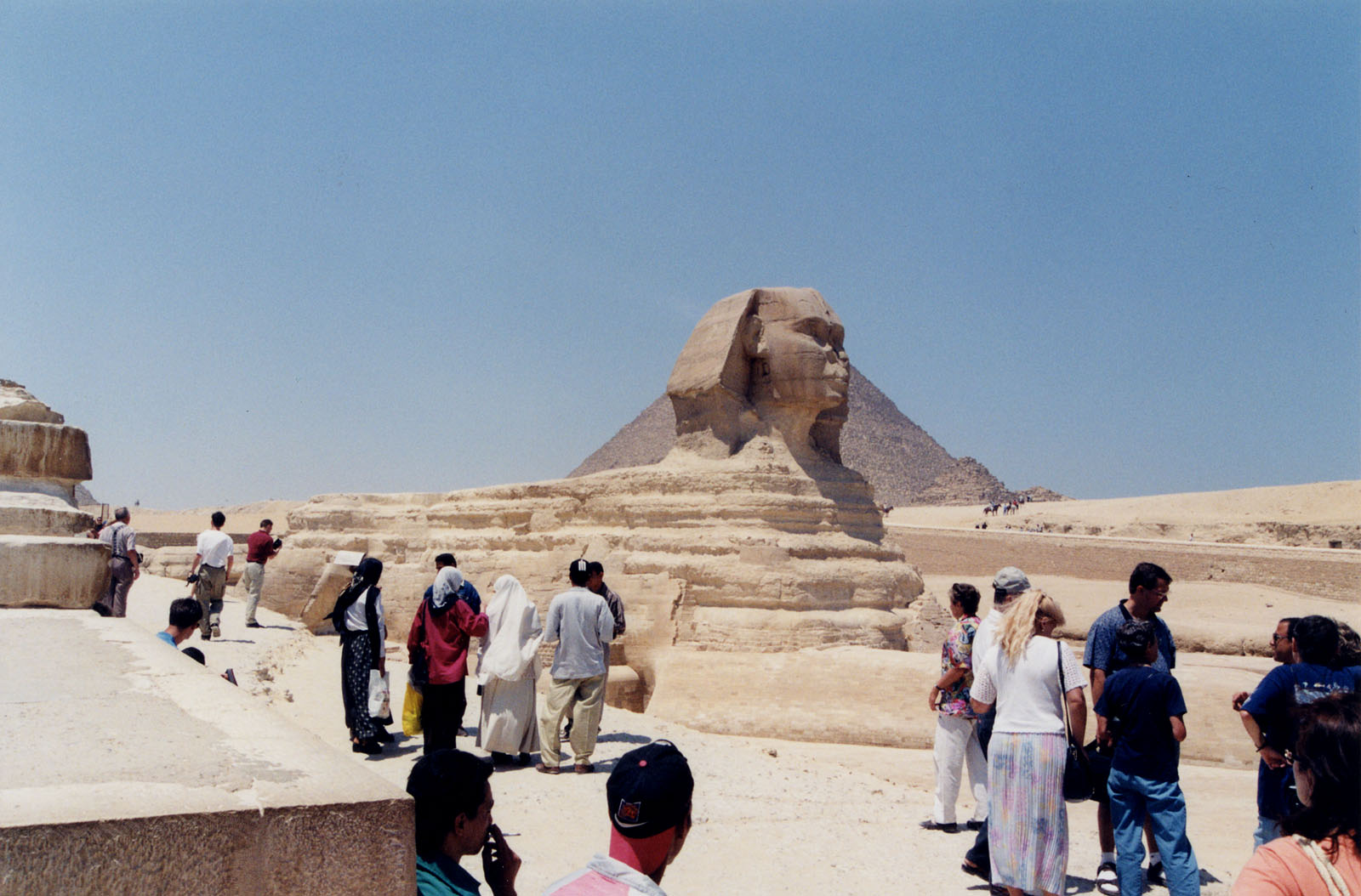 Color photo of the Sphinx from afar with a pyramid in the background and several visitors walking in the foreground